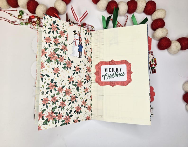 May include: Open journal with pages featuring poinsettia floral patterns and lined paper. A sticker with a snowman and child is on the left page. The right page has a "Merry Christmas" sticker. Red and white felt ball garland decorates the background.