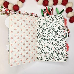 May include: Open journal with patterned pages. One side features a cream background with red floral designs, the other with a white background and green holly leaves. Red and green ribbons mark pages. A festive garland of red and white balls decorates the background.
