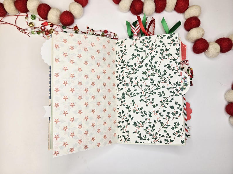 May include: Open journal with patterned pages. One side features a cream background with red floral designs, the other with a white background and green holly leaves. Red and green ribbons mark pages. A festive garland of red and white balls decorates the background.