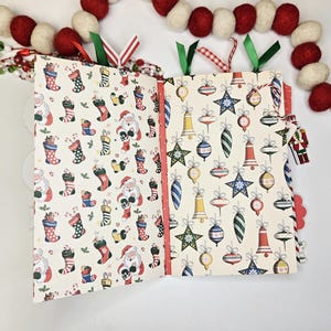 May include: Open Christmas-themed scrapbook with patterned pages. One side features Santa Claus and stockings, the other side has ornaments. Red and green ribbons mark the pages. A garland of red and white felt balls decorates the top.