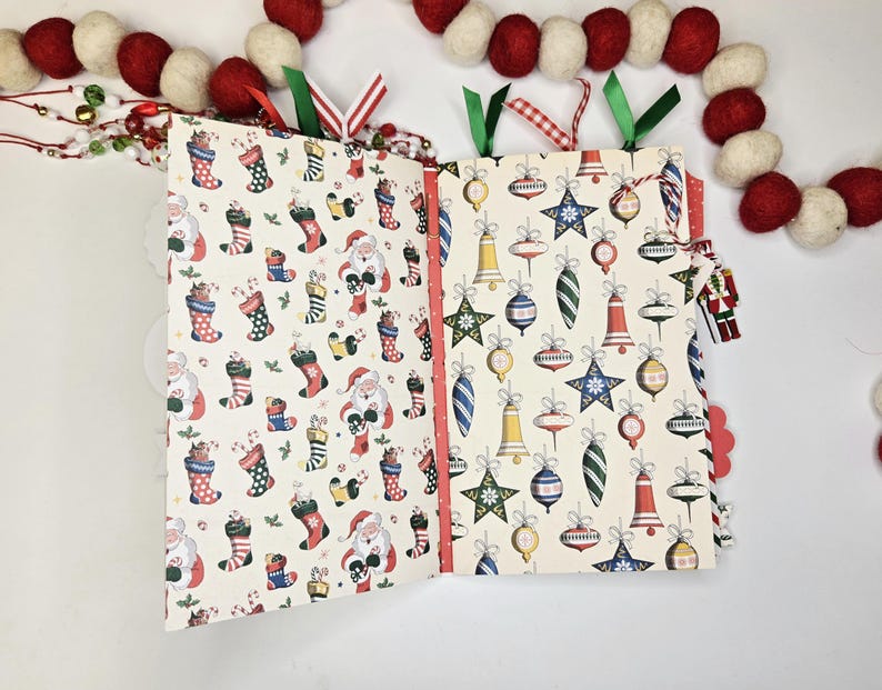 May include: Open Christmas-themed scrapbook with patterned pages. One side features Santa Claus and stockings, the other side has ornaments. Red and green ribbons mark the pages. A garland of red and white felt balls decorates the top.