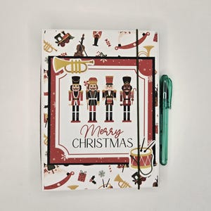 May include: A rectangular notebook featuring a white cover with a pattern of nutcrackers, trains, and musical instruments. The center displays four nutcracker figures and the text Merry CHRISTMAS. A black elastic band and a green pen are attached.