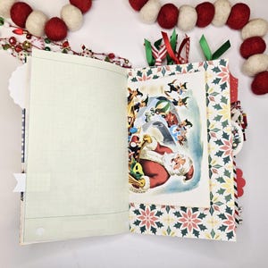 May include: Open journal with a Christmas theme. The journal features a vintage illustration of Santa Claus surrounded by elves. The pages have a grid pattern, and the cover has a festive holly and berry design. Red and white felt ball garland decorates the background.