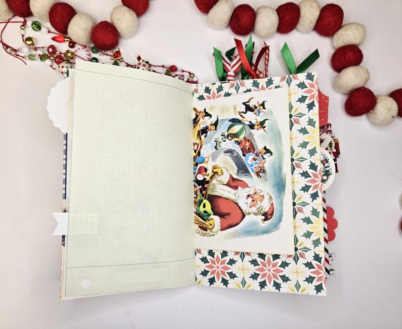 May include: Open journal with a Christmas theme. The journal features a vintage illustration of Santa Claus surrounded by elves. The pages have a grid pattern, and the cover has a festive holly and berry design. Red and white felt ball garland decorates the background.