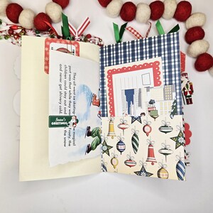 May include: A handmade Christmas-themed journal with a cream-colored cover and patterned interior pages. The journal features a blue and white gingham pocket, a page with a vintage illustration, and a decorative garland.