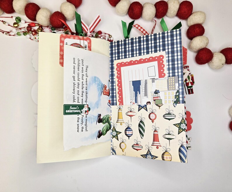 May include: A handmade Christmas-themed journal with a cream-colored cover and patterned interior pages. The journal features a blue and white gingham pocket, a page with a vintage illustration, and a decorative garland.