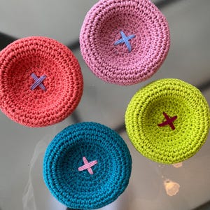 May include: Four circular, bowl-shaped crochet items in pink, coral, lime green, and teal. Each piece features a contrasting X-shaped stitch in the center, with colors including light blue, purple, pink, and dark magenta respectively.