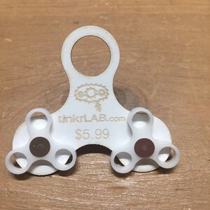 May include: A white fidget spinner with three-pronged arms and a central bearing. The spinner has a circular handle and the text "tinkrLAB.com" is printed on the body. The central bearings are brown.