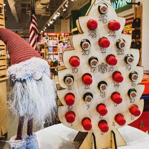 May include: A wooden Christmas tree-shaped shot glass holder with 25 numbered slots. The slots are filled with red shot glasses. The holder is standing on a table with a white surface. The holder is decorated with a star at the top and the word "SHOTS" written on the star.