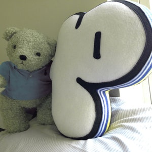 Op de afbeelding: Een pluche grijze teddybeer met een blauw poloshirt zit naast een groot wit kussen met zwarte en blauwe strepen. Het kussen is gevormd als de letter "P" met een zwarte omtrek.