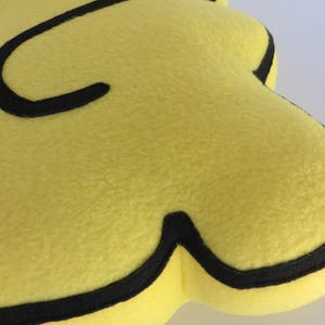 Op de afbeelding: Een geel pluche speelgoed in de vorm van de letter C met een zwarte omtrek. Het speelgoed is gemaakt van zacht, pluche stof.