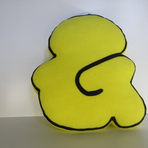 Op de afbeelding: Een geel kussen in de vorm van een ampersand met een zwarte omtrek.