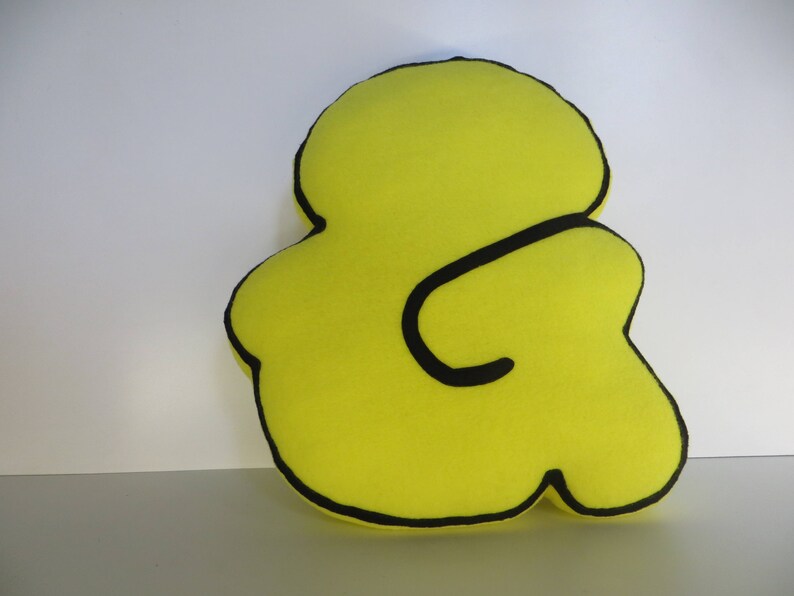 Op de afbeelding: Een geel kussen in de vorm van een ampersand met een zwarte omtrek.