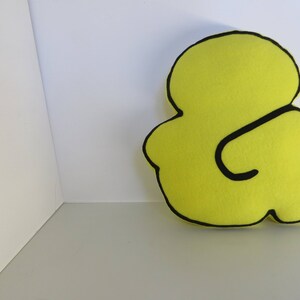 Op de afbeelding: Een geel kussen in de vorm van een ampersand met zwarte contouren.