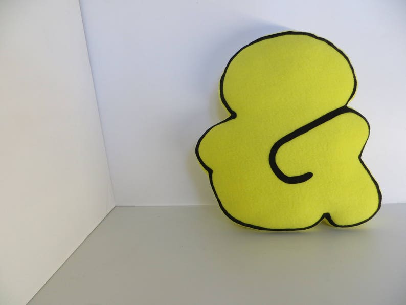 Op de afbeelding: Een geel kussen in de vorm van een ampersand met zwarte contouren.