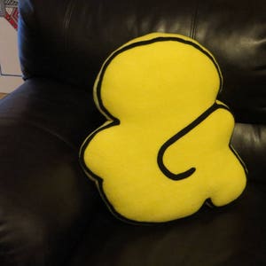 Op de afbeelding: Een geel kussen in de vorm van een ampersand met een zwarte omtrek. Het kussen ligt op een zwarte leren bank.