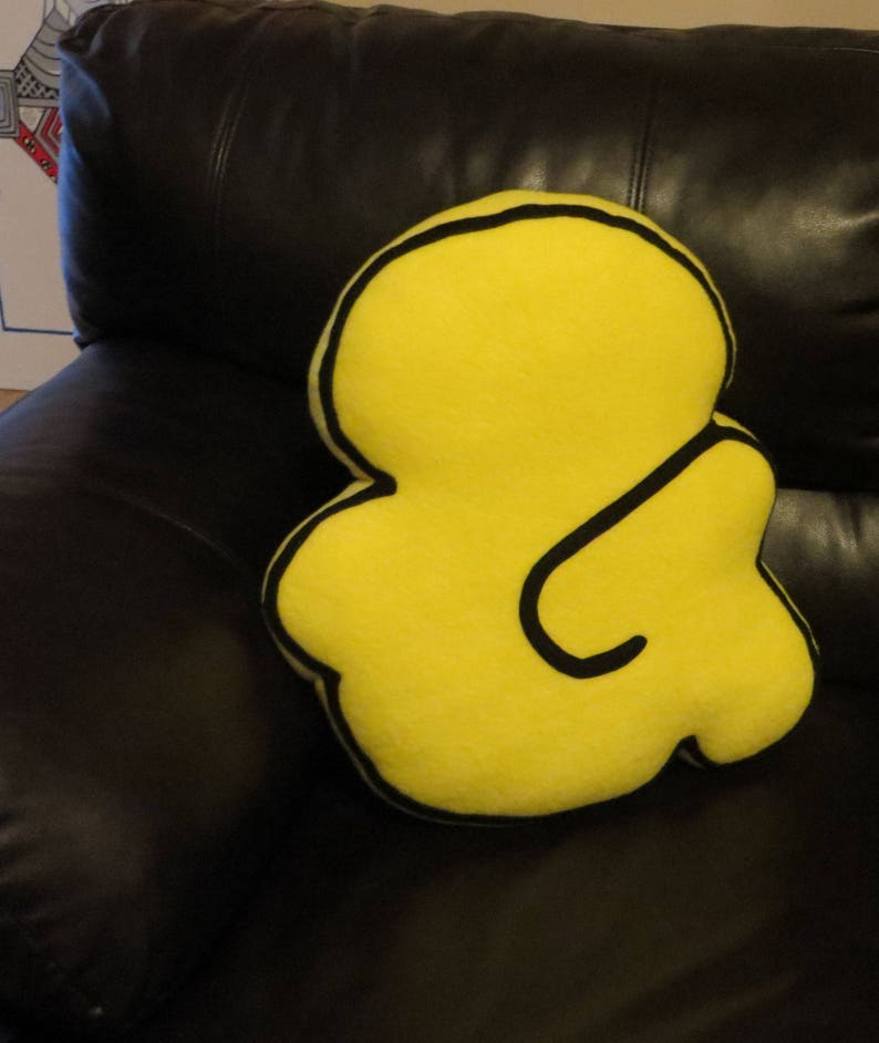 Op de afbeelding: Een geel kussen in de vorm van een ampersand met een zwarte omtrek. Het kussen ligt op een zwarte leren bank.