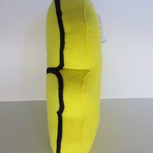 Op de afbeelding: Een geel pluche kussen met een zwarte omtrek in de vorm van een banaan.