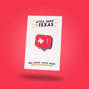 Puede incluir: Un pin de esmalte rojo y dorado con un contorno blanco del estado de Texas dentro de una forma de corazón rojo con borde dorado. El pin tiene un número "1" dorado al lado del corazón. El pin está en una tarjeta blanca con el texto "LITTLE SHOP OF TEXAS" en letras doradas y el texto "BIG STATE. LITTLE SHOP. WWW.LITTLESHOPOFTX.COM" en letras negras.