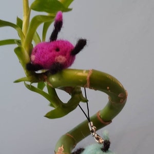 May include: Two small, felt, plush toys hanging from a bamboo plant. The top toy is pink with black accents and the bottom toy is blue with black accents.