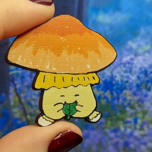 May include: A hand-painted wooden mushroom figurine with a yellow cap and a smiling face. The mushroom is holding a green leaf in its hands.