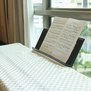 May include: A black music stand with sheet music on a white and gray checkered table top. The music stand is in front of a window with a view of a city.