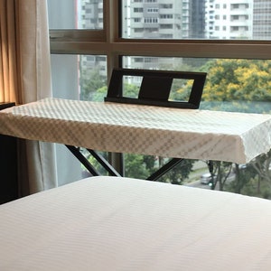 May include: White keyboard cover with a checkered pattern, sitting on a black keyboard stand. The stand is positioned over a white bed with a window in the background.