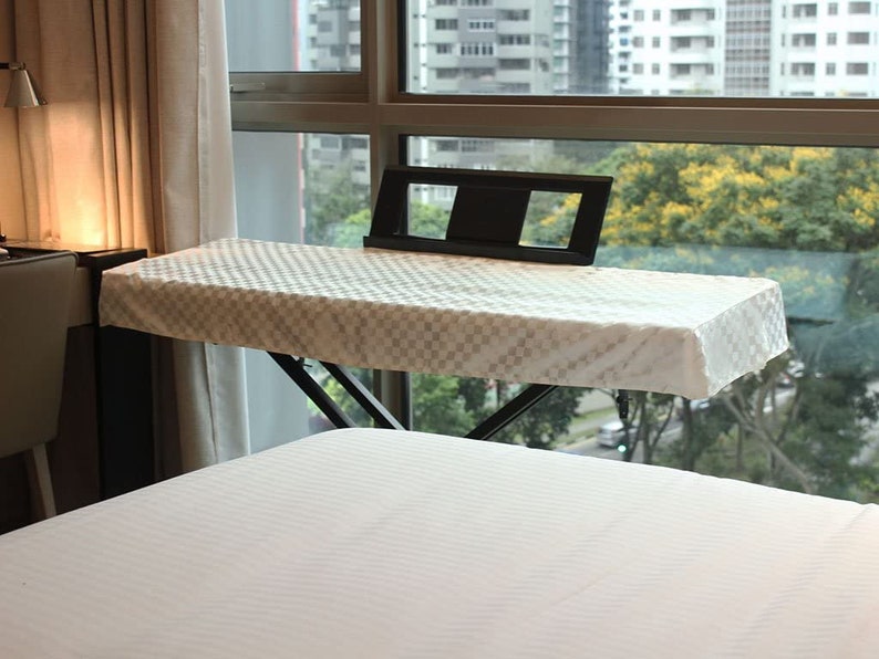 May include: White keyboard cover with a checkered pattern, sitting on a black keyboard stand. The stand is positioned over a white bed with a window in the background.