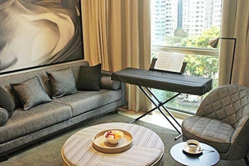 May include: A black keyboard stand with a black keyboard cover. The stand is in a living room with a gray couch, a gray armchair, and a coffee table with a bowl of fruit.