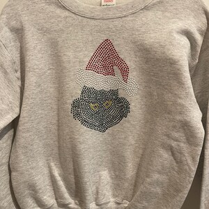 May include: A light gray sweatshirt featuring a rhinestone design of a Grinch-like face wearing a Santa hat. The hat is red with white trim, and the face is black with yellow eyes. The sweatshirt is a crew neck style.