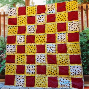 Op de afbeelding: Een handgemaakte patchworkquilt met een rasterpatroon. De quilt heeft afwisselende vierkanten van effen bordeauxrood, geel met zwarte stippen en crème met bruine hartpatronen. De randen hebben een gerafelde, getextureerde afwerking.