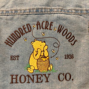 Könnte beinhalten: Hellblaue Jeansjacke mit einem gestickten Design. Es zeigt einen gelben Bären, einen Honigtopf und den Text "Hundred Acre Woods Honey Co. Est. 1926". Das Design umfasst Bienen und ist mit braunem Faden gestickt.