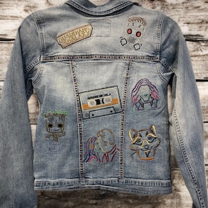 Könnte beinhalten: Hellblaue Jeansjacke mit aufgestickten Aufnähern mit Charakteren und Text aus Guardians of the Galaxy. Zu den Aufnähern gehören Star-Lord, Groot, Rocket und eine Kassette. Die Jacke hat einen klassischen Kragen und einen Knopfverschluss.