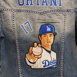 Könnte beinhalten: Jeansjacke mit dem Namen "Ohtani" in Blau auf dem Rücken gestickt. Die Nummer "17" ist ebenfalls auf die Jacke gestickt. Ein Aufnäher eines Baseballspielers im Dodgers-Trikot befindet sich auf der Jacke.