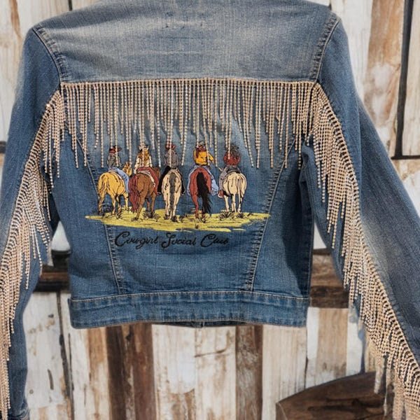 May include: A light blue denim jacket with a rhinestone fringe along the back and sleeves. The back of the jacket features an embroidered design of several people on horseback with the text "Cowgirl Social Club".