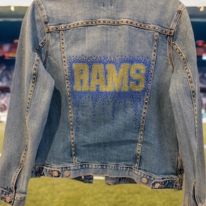 Könnte beinhalten: Hellblaue Jeansjacke mit dem Wort "RAMS" aus goldenen und blauen Strasssteinen auf dem Rücken. Die Jacke hat ein klassisches Design mit Knopfverschlüssen und Taschen und ist mit goldenen Strasssteinen entlang der Nähte verziert.