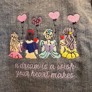 May include: Embroidered denim jacket featuring four cartoon princesses holding heart-shaped balloons. The text "a dream is a wish your heart makes" is embroidered in purple thread below the figures. The jacket is a medium wash denim.