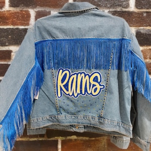 Può includere: Giacca di jeans azzurra con frange blu brillante e la scritta "Rams" in giallo. La giacca ha un colletto classico e una chiusura con bottoni. Piccoli abbellimenti color oro sono sparsi intorno alle lettere.