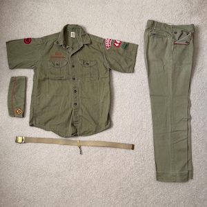 May include: An olive green Boy Scout uniform set, including a short-sleeved shirt with patches, matching pants, a belt, and a shoulder sleeve insignia. The shirt has two chest pockets and button closures. The pants have red trim.