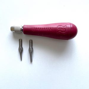 May include: A Speedball calligraphy pen set with a burgundy handle and two nibs. The handle has the word "SPEEDBALL" printed on it. The nibs are silver and have a pointed tip. The pen set is designed for calligraphy and lettering.