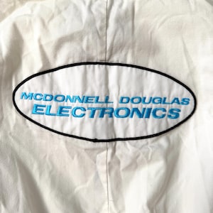 May include: White fabric with an embroidered patch. The patch is an oval shape with a black border and the words "McDonnell Douglas Electronics" in blue.