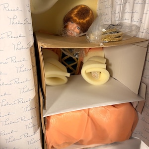 May include: A vintage doll in its original packaging. The doll has red hair and is wearing a dress with green and gold accents. The packaging includes tissue paper and foam supports to protect the doll.