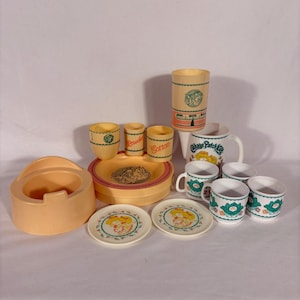 May include: A collection of vintage Cabbage Patch Kids toy dishes and accessories in shades of peach, yellow, and white. Includes plates, cups, a pitcher, and a container labeled "Cotton". The set features the Cabbage Patch Kids logo and graphics.