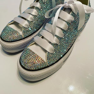 Puede incluir: Un par de zapatillas de lona blancas cubiertas de pedrería iridiscente con cordones de satén blanco.