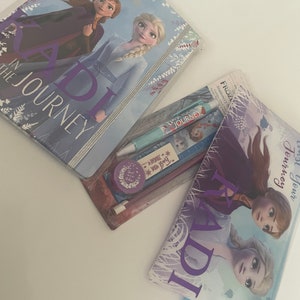 Peut inclure: Une trousse à crayons violette avec un motif Frozen 2 représentant Anna et Elsa. La trousse porte le texte "KADI" en grandes lettres violettes. La trousse est ouverte et montre un jeu de crayons avec un motif Frozen 2.