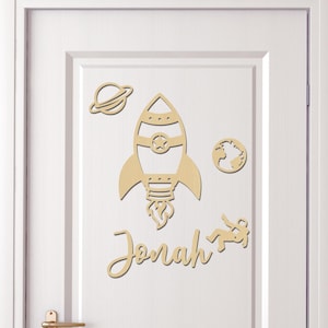 Può includere: Decorazione per porta in legno con sagome di razzo, pianeti e astronauta. Il nome "Jonah" è scritto in corsivo.