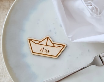 May include: A wooden paper boat place card with the name Nils written on it. The place card is on a white plate with a white napkin.