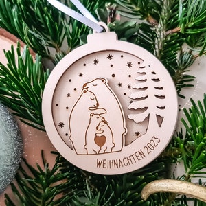 May include: A wooden Christmas ornament shaped like a bauble with a cutout design of a family of polar bears hugging. The ornament is decorated with snowflakes and a small pine tree. The text "WEIHNACHTEN 2023" is inscribed on the bottom.
