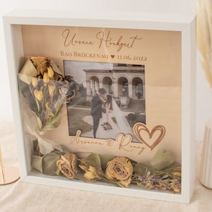 Può includere: Una cornice bianca a scatola d'ombra con una foto di matrimonio all'interno. La foto mostra una sposa e uno sposo davanti a un edificio. La cornice contiene anche fiori secchi e un cuore con le parole "Unsere Hochzeit" e "Bad Brückenau" scritte su di essa.