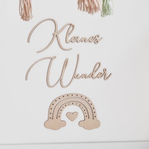May include: Wooden wall decor with the words "Kleines Wunder" in cursive script, a rainbow with a heart in the center, and a white background.